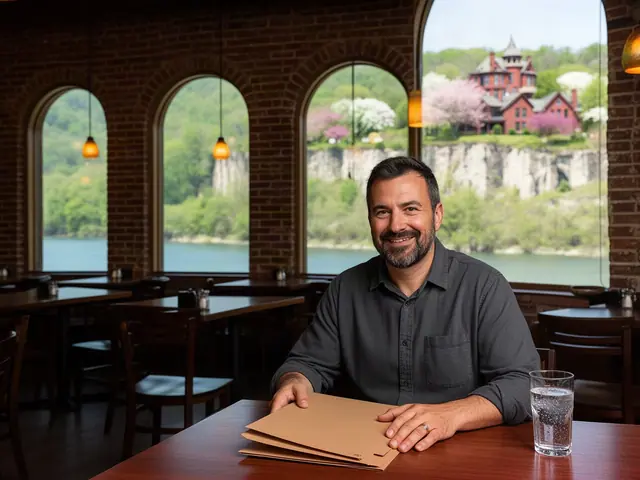 Smiling bistro owner in a charcoal shirt at a table with closed folders, arched windows onto bluffs and the river